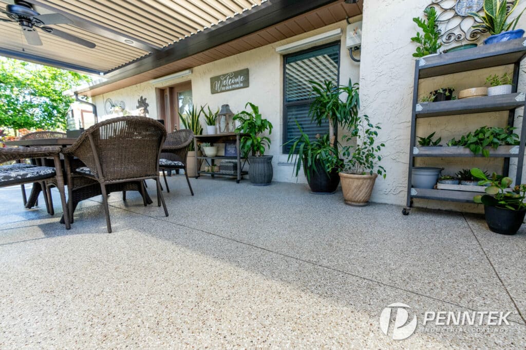 Beautifully coated covered porch with durable, speckled concrete finish by Penntek Industrial Coatings, showcasing a welcoming seating area, lush green plants, and rustic decor elements.